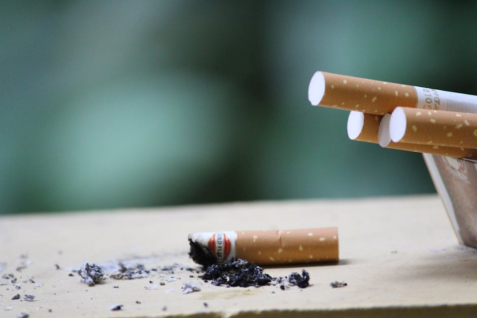 Close-up of a broken cigarette with burnt ash and debris scattered on a light wooden surface, with five intact cigarettes in the background. The scene is illuminated by soft natural light, and the bac