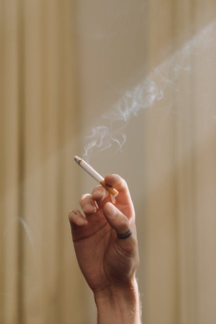 Close-up of a broken cigarette with burnt ash and debris scattered on a light wooden surface, with five intact cigarettes in the background. The scene is illuminated by soft natural light, and the bac