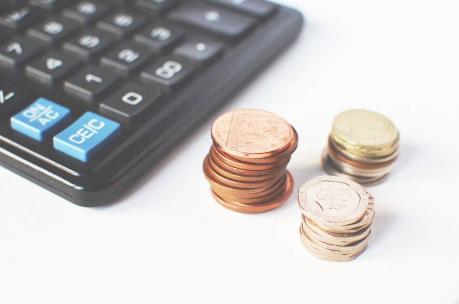Close-up image of a calculator with a black body and light blue function keys, placed on a white surface. Next to the calculator are three neat stacks of Euro coins, with the largest stack comprising 